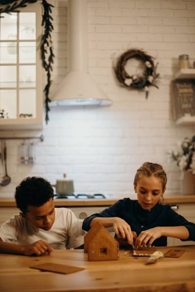 Two kids building a gingerbread house at the kitchen table