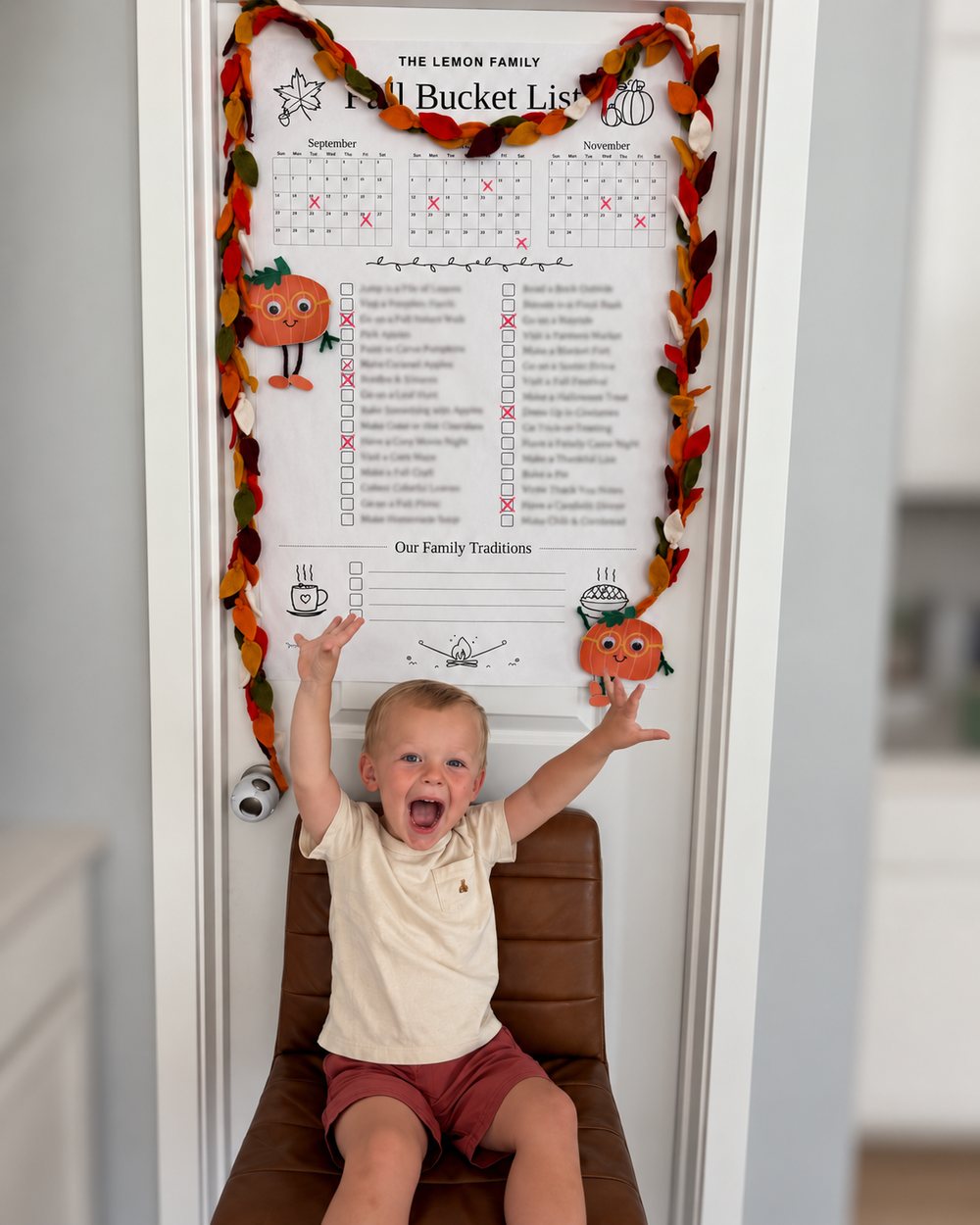 A toddler cheering with both arms up in front of the Smith Family Fall Bucket List calendar on a door